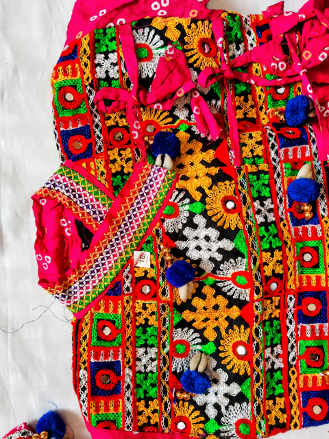 A vibrant, heavily embroidered traditional Gujrati Kedia laid flat on a white fabric background. The garment features intricate multicolor embroidery with prominent pink, green, yellow, orange, and black patterns, decorated with blue pom-poms and small wooden beads.