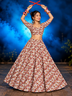 A woman modeling a floral print lehenga choli set, holding red decorative sticks above her head against a smoky blue studio background.